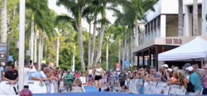 A woman athlete running down the pole vault runway with a large crowd at an outdoor event in a Naples plaza