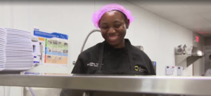 A smiling St. Matthew's House cook wearing a black chef's coat and pink hair net working in a commercial kitchen.
