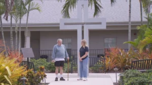 Two people having a conversation outdoors in a courtyard in front of a large cross, surrounded by tropical landscaping.